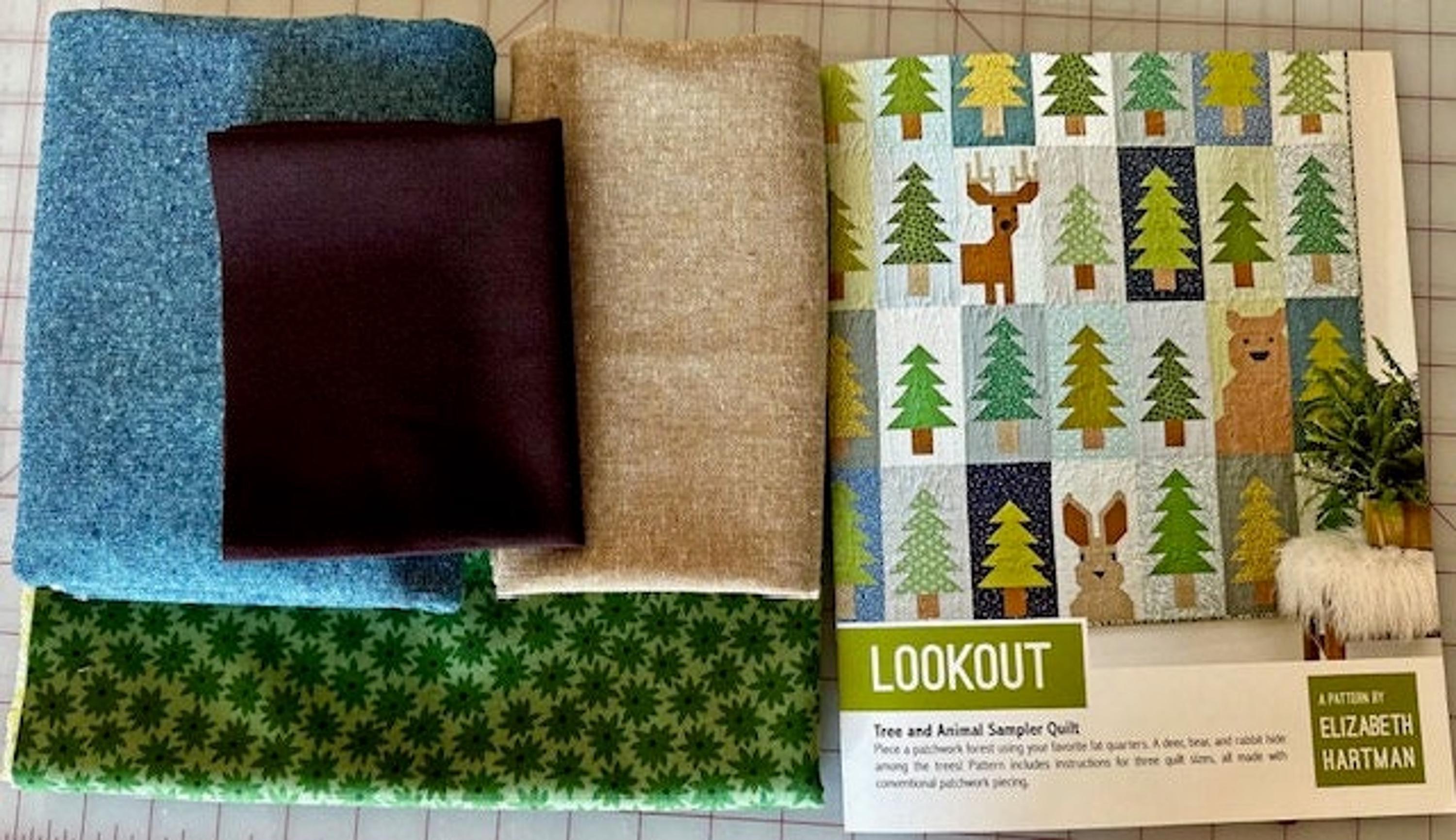 Four folded fabric pieces in blue, brown, and tan, next to a quilt pattern booklet featuring a quilt with trees and animals.