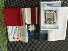 Various folded fabrics, including red, blue, and white star patterns, and a quilt pattern booklet, are arranged on a black cutting mat.