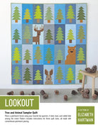 A finished quilt with a grid pattern of green pine trees and brown forest animals, displayed on a white surface with pinecones scattered around.